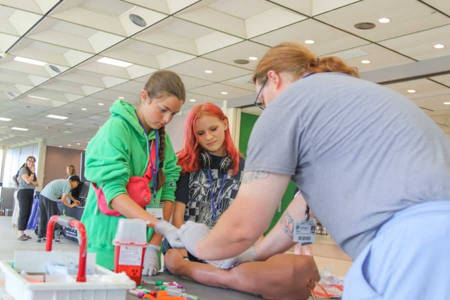 Two young students looking over an adult simulating a medical test on a manequeen arm/ 