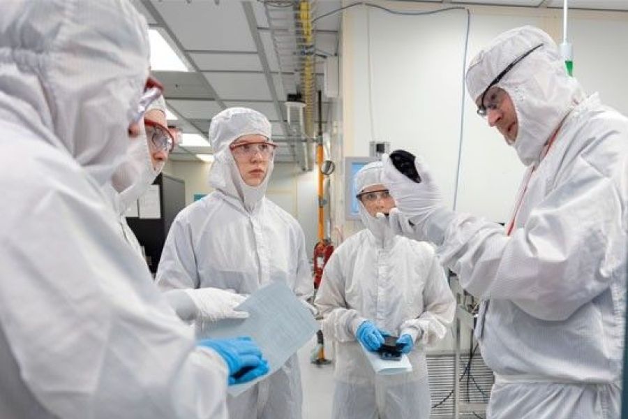Group of people wearing cleanroom suits and safety goggles in a laboratory setting, with one person holding up a device for inspection.