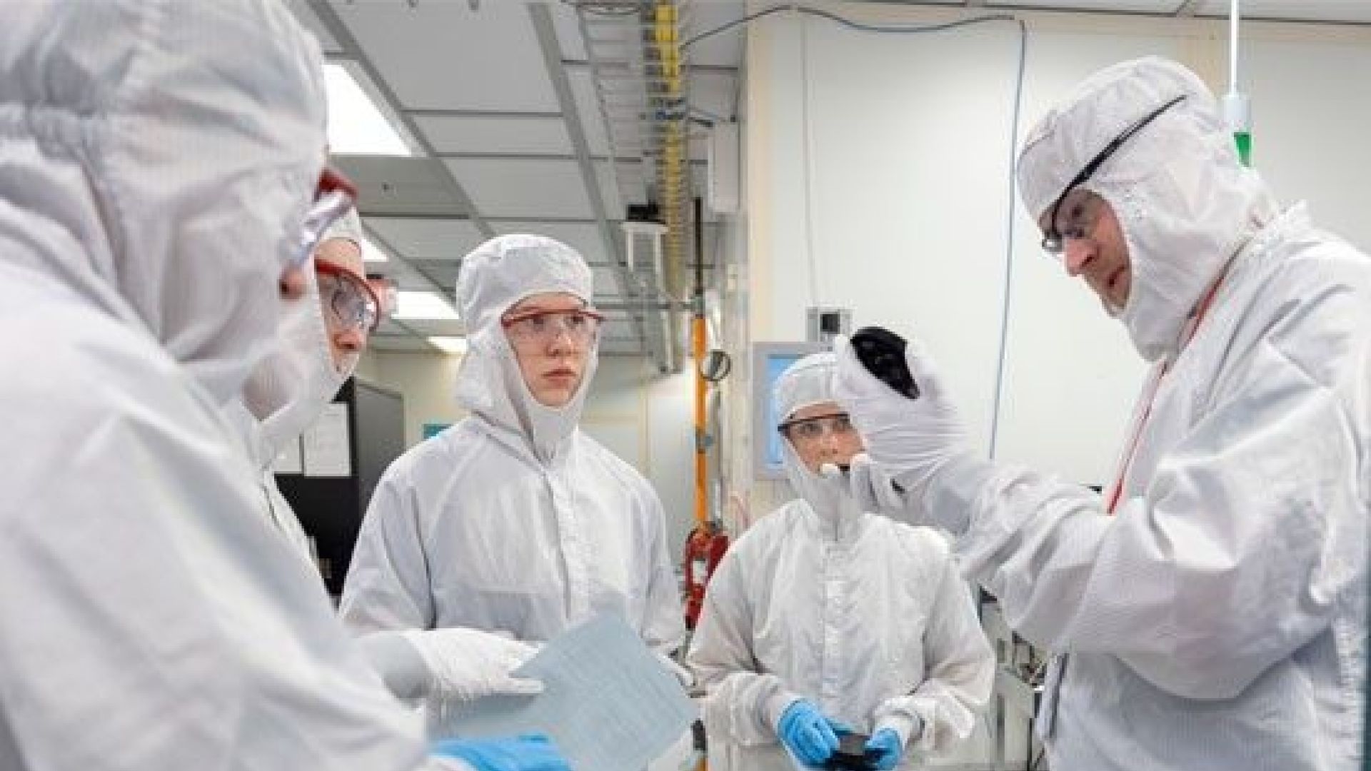Group of people wearing cleanroom suits and safety goggles in a laboratory setting, with one person holding up a device for inspection.